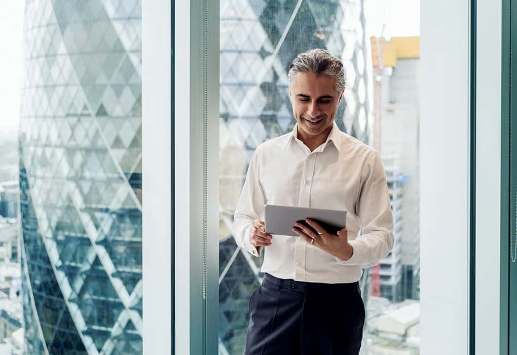 A man in a white shirt stands by a large window, looking at a tablet. Modern glass buildings are visible outside the window.