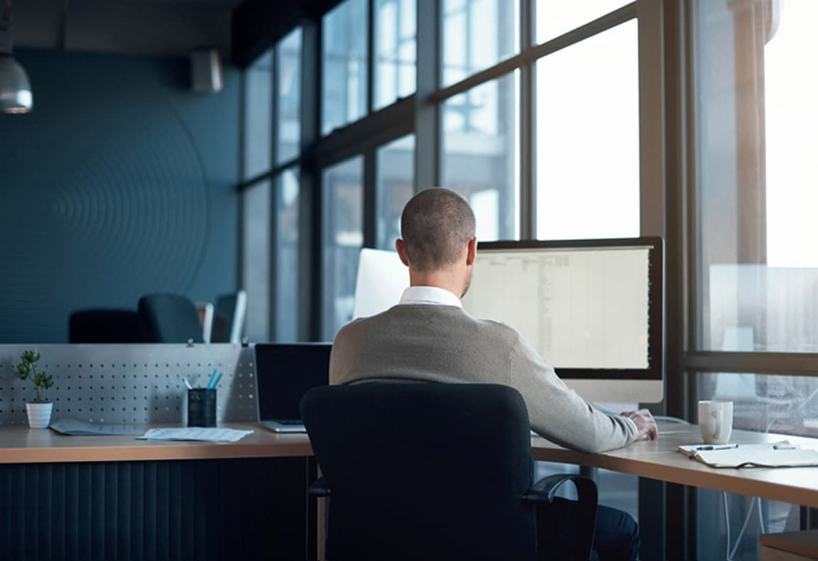 A person sitting at a desk in an office, viewed from behind, working on a computer with large windows in the background.