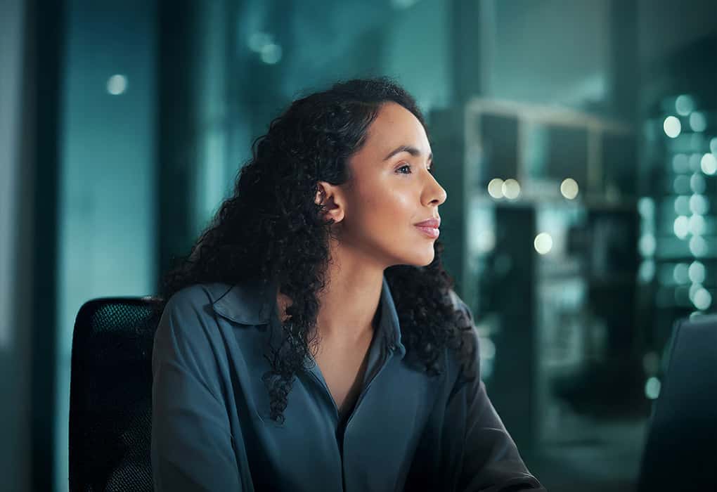 A woman with curly hair, wearing a dark shirt, is looking to the side while sitting in an office setting with soft lighting.