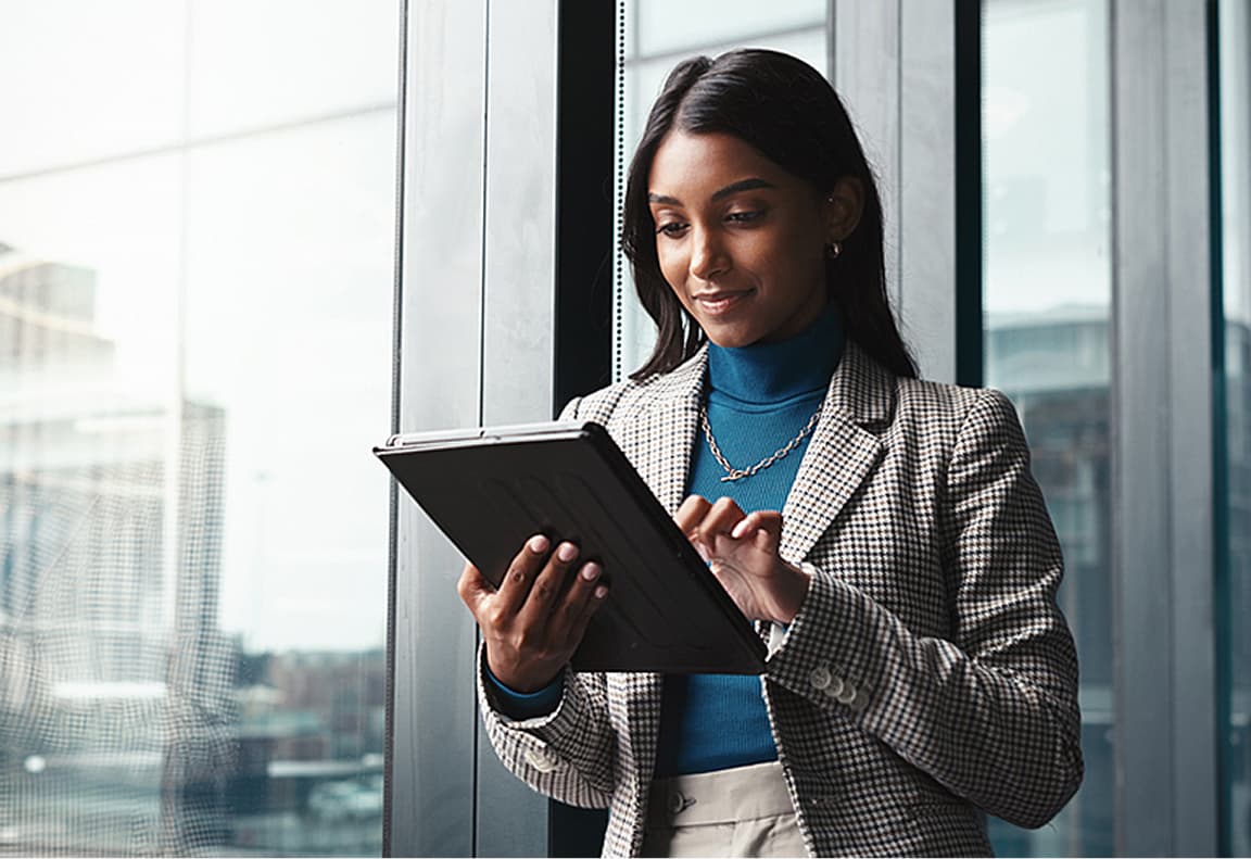 A young woman in a checked blazer and blue turtleneck is using a tablet while standing by a large window with an urban view in the background.