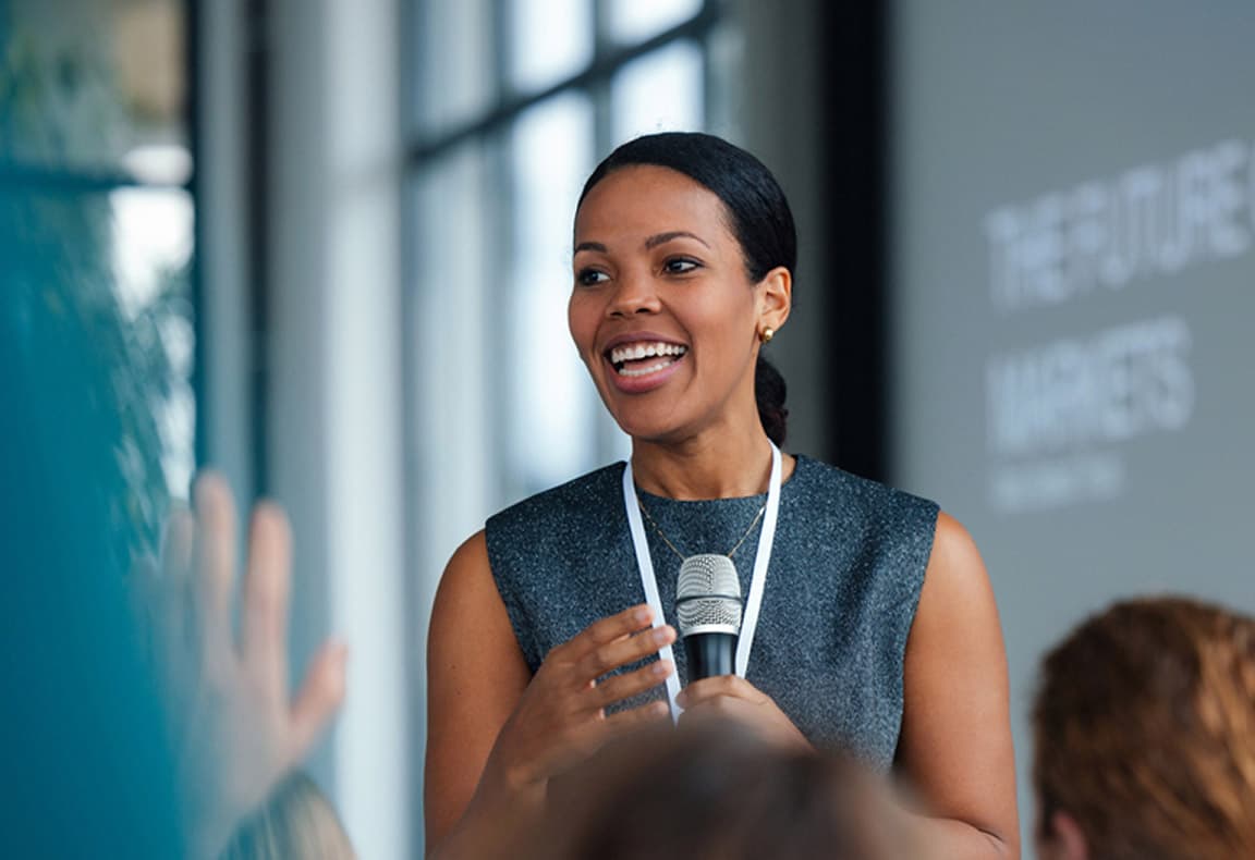 A woman with dark hair, wearing a sleeveless gray dress, is speaking into a microphone while smiling at the audience in a bright, modern setting.